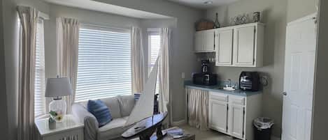 Ein gemütlicher Wohnbereich mit Küchenzeile, Deckenventilator, einer Couch mit Kissen, einem kleinen Tisch und einem Fenster mit Vorhängen.