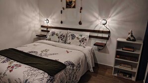 Una camera da letto con una coperta a fiori, una testiera in legno, lampade sui comodini, una mensola con libri e un quadro appeso al muro.