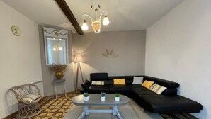 Un salon avec un canapé d’angle noir, une table basse ronde agrémentée de plantes, un fauteuil en osier, un lampadaire et un tapis à motifs.
