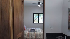 Ein schmaler Raum mit Bett, Fenster, Deckenventilator und Bad mit Waschbecken und Handtuch.