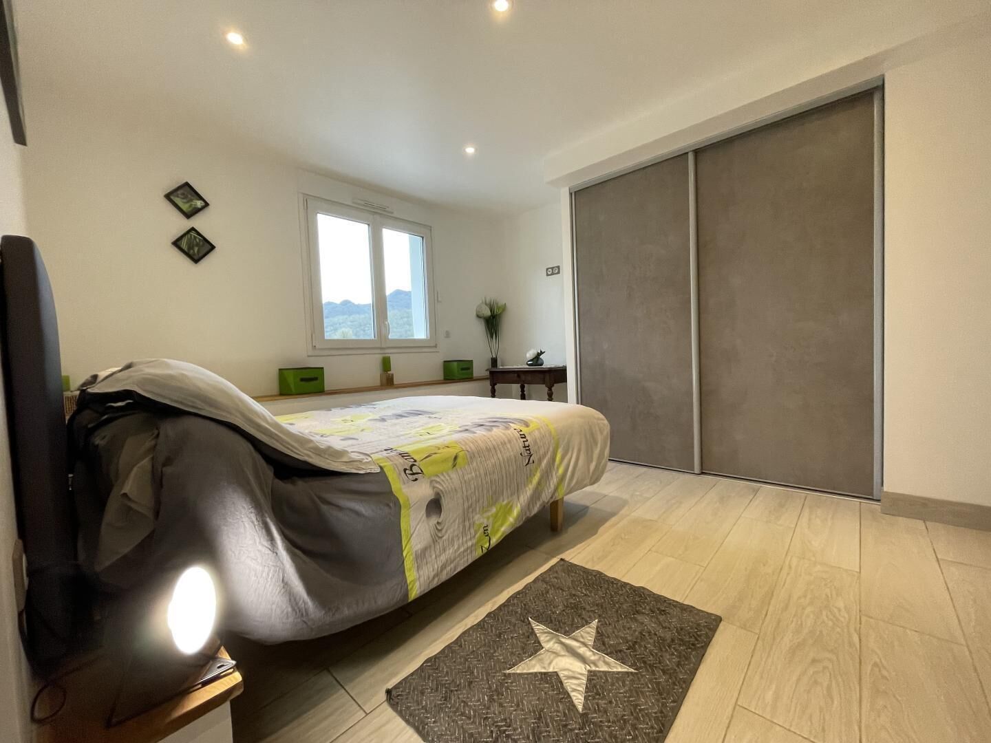 Une chambre à coucher avec un lit recouvert d’une couverture, une table de chevet avec une lampe, une fenêtre donnant sur un paysage agréable et une porte coulissante.