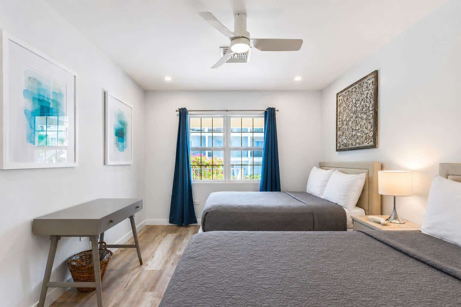 Habitación de hotel con dos camas, un escritorio, ventilador de techo, ventana con cortinas y obras de arte en las paredes.