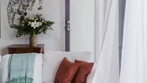 A white sofa with a teal throw blanket and two brown pillows, a small wooden side table with a vase of flowers, and a painting on the wall.