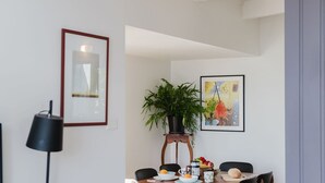 A dining area with a table set for two, a potted plant, and a framed artwork on the wall.