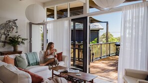 A spacious living area with a large window, a sofa, a coffee table, and a view of the outdoor deck. - A Little Fish in Mollymook Beach (Mollymook)