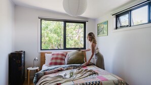 A bedroom with a large window, a bed with multiple pillows, a bedside table with a lamp, and a suitcase.