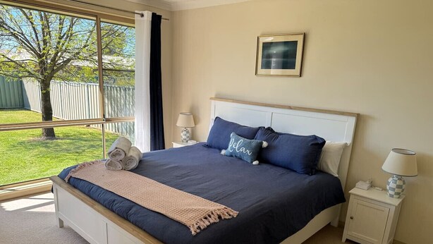 A bedroom with a large bed, a nightstand, a lamp, a framed picture on the wall, and a view of a green lawn and tree through the window.