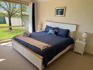 Ein Schlafzimmer mit einem großen Bett, einem Nachttisch, einer Lampe, einem gerahmten Bild an der Wand und Blick auf eine grüne Wiese und einen Baum durch das Fenster.
