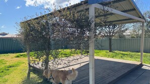 A covered outdoor area with a wooden deck, surrounded by a grassy yard and a metal fence.
