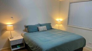 A bedroom with a bed, nightstands, lamps, and a window with blinds. - Spacious & comfy studio unit “Rest Easy Mudgee” (Mudgee)