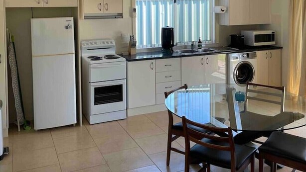 A compact kitchen with white appliances, a dining table with chairs, and a window with blinds. - Spacious & comfy studio unit “Rest Easy Mudgee” (Mudgee)