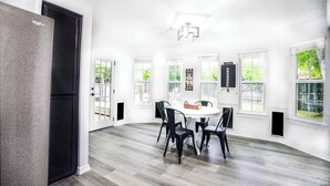 Ett rymligt matområde med ett runt bord och fyra stolar, stora fönster och ett modernt kök som syns i bakgrunden.
