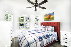 Ein Schlafzimmer mit einem großen Bett, einem roten Kopfteil, einem Deckenventilator und einem Fenster mit Blick auf Grünflächen.