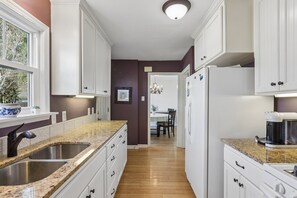 A kitchen with white cabinets, granite countertops, a double sink, and a refrigerator. - Cozy 2-Bedroom Brookside Bungalow in Kansas City - Walkable to Shops and Dining (Kansas City)