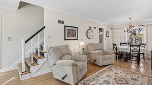 Ein Wohnzimmer mit Treppe, zwei beigefarbene Sessel, einem Esstisch mit Stühlen und einer Chandelüster.