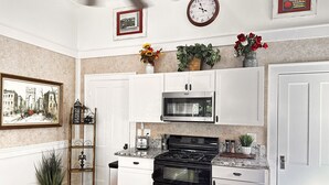A compact kitchen with white cabinets, a black stove, a microwave, and a small oven. There are decorative plants and a framed picture on the wall.