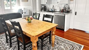 A dining area with a wooden table and chairs, a kitchen with white cabinets, and a rug on the floor.