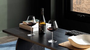 Une table en bois foncé, sur laquelle se trouvent une bouteille de vin et deux verres, un livre et un clavier.