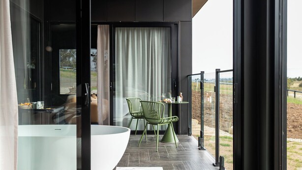 Une chambre moderne avec un grand lit, une porte vitrée donnant sur un balcon équipé d’un coin repas et offrant une vue sur l’extérieur.