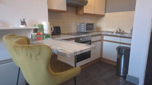 Een kleine keuken met een magnetron, fornuis en wastafel. Twee gele stoelen en een tafeltje staan er.