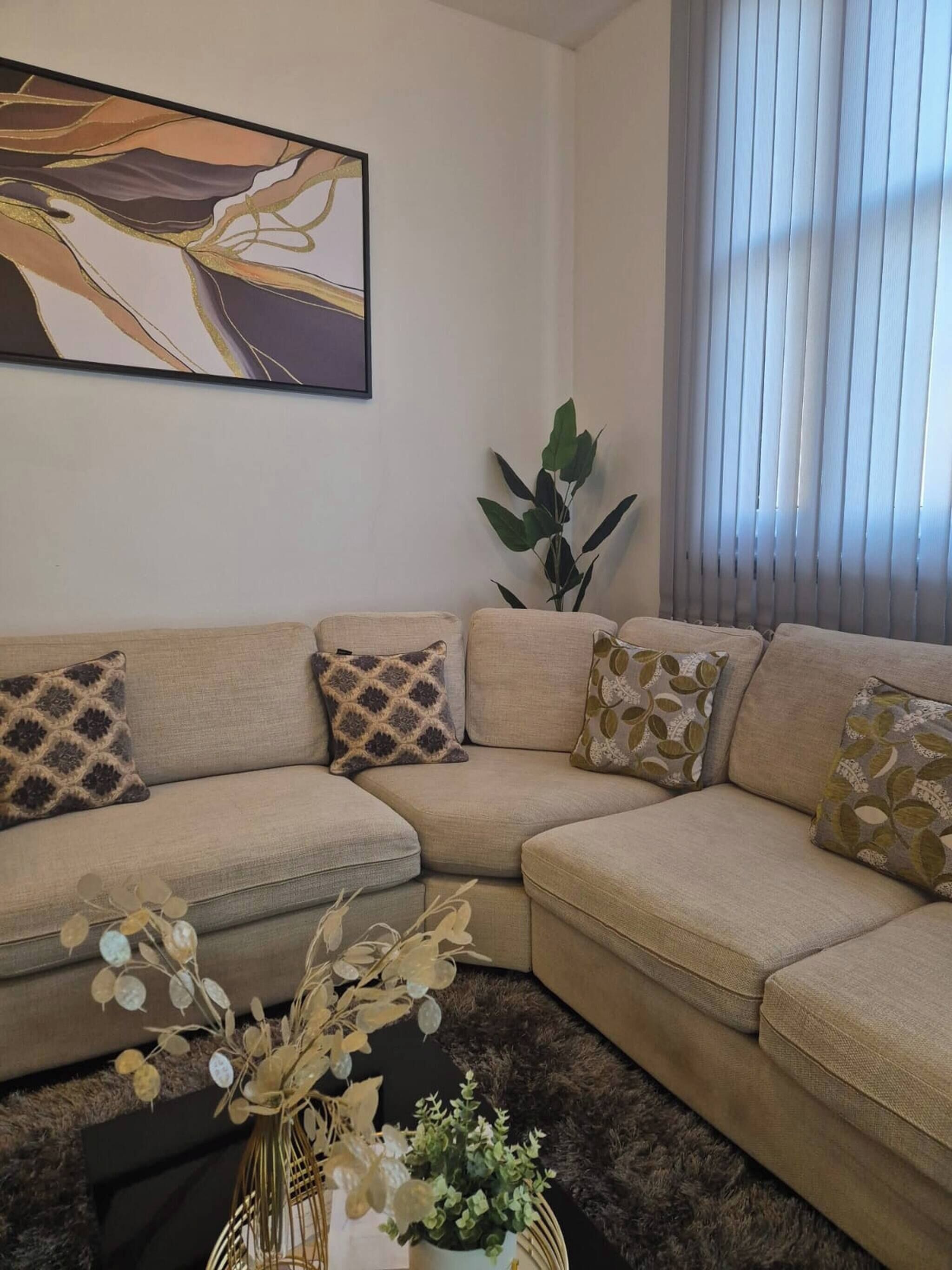 A living room with a beige sectional sofa, patterned cushions, a large abstract painting, a potted plant, and a window with blinds.