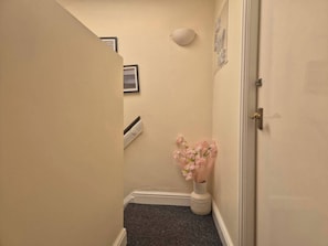 Un pasillo estrecho con una puerta a la derecha, un jarrón pequeño con flores rosas en el suelo y una lámpara fijada a la pared.