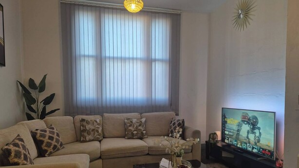 Een woonkamer met een modulaire bank, een salontafel met een vaas, een televisie op een meubelstuk, en een groot raam met jaloezieën.