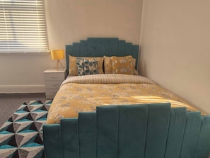 Un dormitorio con una cama tapizada en color aguamarina, ropa de cama floral, un tapete con estampado geométrico, una mesita de noche con lámpara y una ventana con persianas.