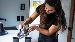 Une personne verse du café dans deux tasses sur un plan de travail blanc, dans une cuisine moderne dotée d’une fenêtre et d’une porte.