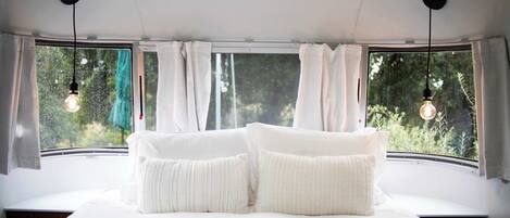 A bedroom with a large bed, two hanging light bulbs, and a view of greenery through the window.