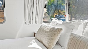 Un lit avec une literie blanche et un oreiller à texture, une fenêtre donnant sur l’extérieur et une ampoule suspendue.