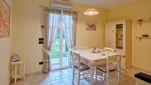 Uma área de refeições com uma mesa e cadeiras, um armário com prateleiras e uma pequena mesa lateral.