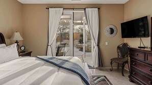 A bedroom with a bed, a TV, a chair, a dresser, and a sliding glass door to a patio. - Blue Jay by Nest Villas | Idyllic Pool & Spa (Indio)