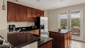 A modern kitchen with dark wood cabinets, a black countertop, stainless steel appliances, and a view of a balcony with outdoor furniture. - Blue Jay by Nest Villas | Idyllic Pool & Spa (Indio)