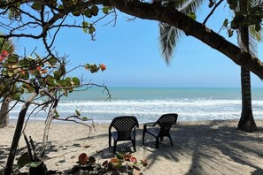 A beach with two black plastic chairs under a palm tree, overlooking the ocean.