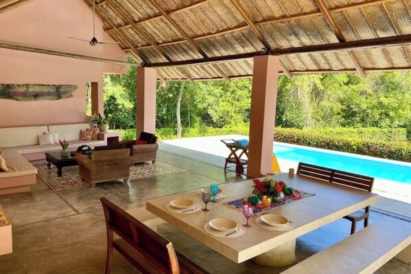 A covered outdoor dining area with a pool in the background.