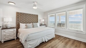 Ein Schlafzimmer mit einem großen Bett, einem Holzkopfteil, zwei Nachttischen mit Lampen und Blick auf das Meer durch große Fenster.