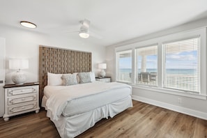 Ein Schlafzimmer mit einem großen Bett, einem Holzkopfteil, zwei Nachttischen mit Lampen und Blick auf das Meer durch große Fenster.