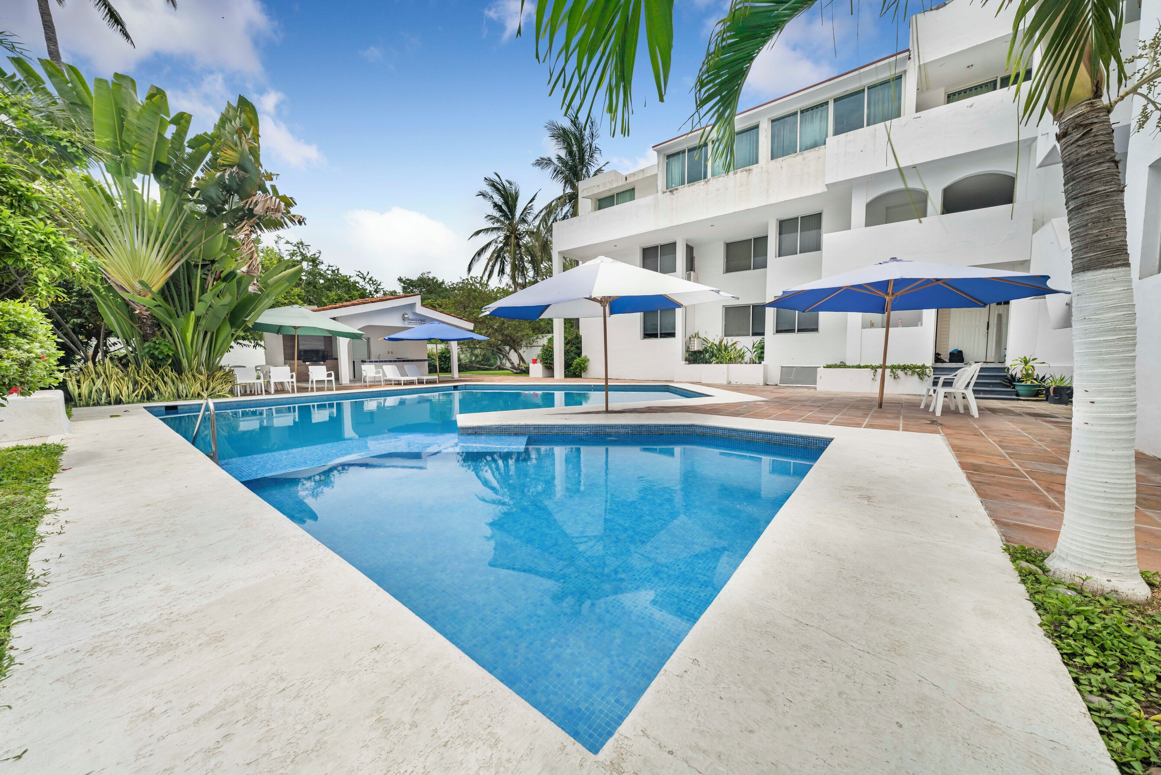 A swimming pool with a white tiled deck, surrounded by palm trees and a multi-story building with balconies.