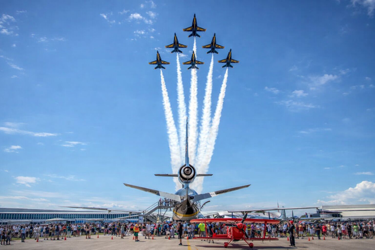 A large crowd gathered at an airshow with a central aircraft performing a stunt.