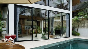 A modern pool area with a glass-enclosed dining space, a wooden table with drinks, and a wicker basket with food. - Ubud Dzen Villas (Ubud)