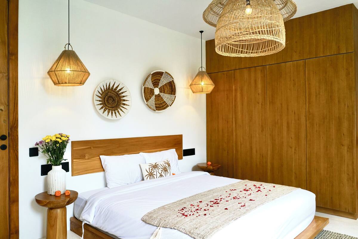 A neatly arranged bedroom with a wooden headboard, a large bed with white bedding, two wall-mounted lights, and a woven pendant light above the bed.