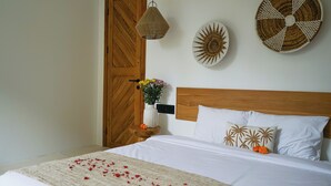 A neatly made bed with white linens, a wooden headboard, and a decorative pillow with palm tree patterns. There is a small wooden nightstand with a vase of flowers and a woven wall hanging. - Ubud Dzen Villas (Ubud)