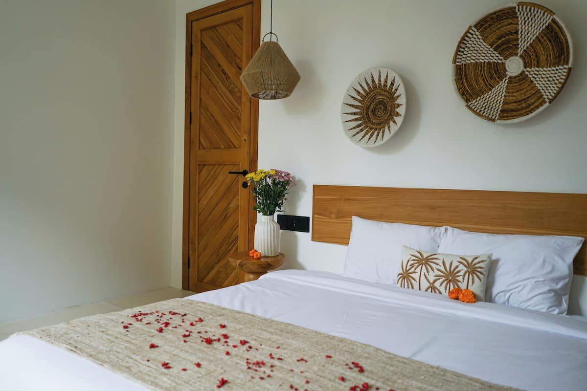 A neatly made bed with white linens, a wooden headboard, and a decorative pillow with palm tree patterns. There is a small wooden nightstand with a vase of flowers and a woven wall hanging.