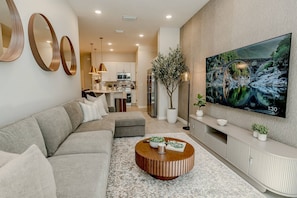 A modern living room with a sectional sofa, a round coffee table, and a large flat-screen TV mounted on the wall. - 5BR Home for 16 w/ Pool & Game Room (Orlando)