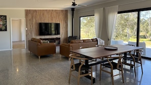 A modern living room with a large TV, a fireplace, a dining area, and large windows. - Modern Luxe Bush Retreat | 3BR Near Trails (Nannup)