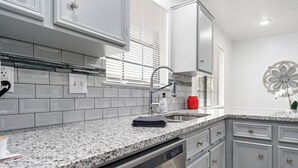 Una cocina con gabinetes blancos, encimeras de granito, lavavajillas y una ventana con persianas.