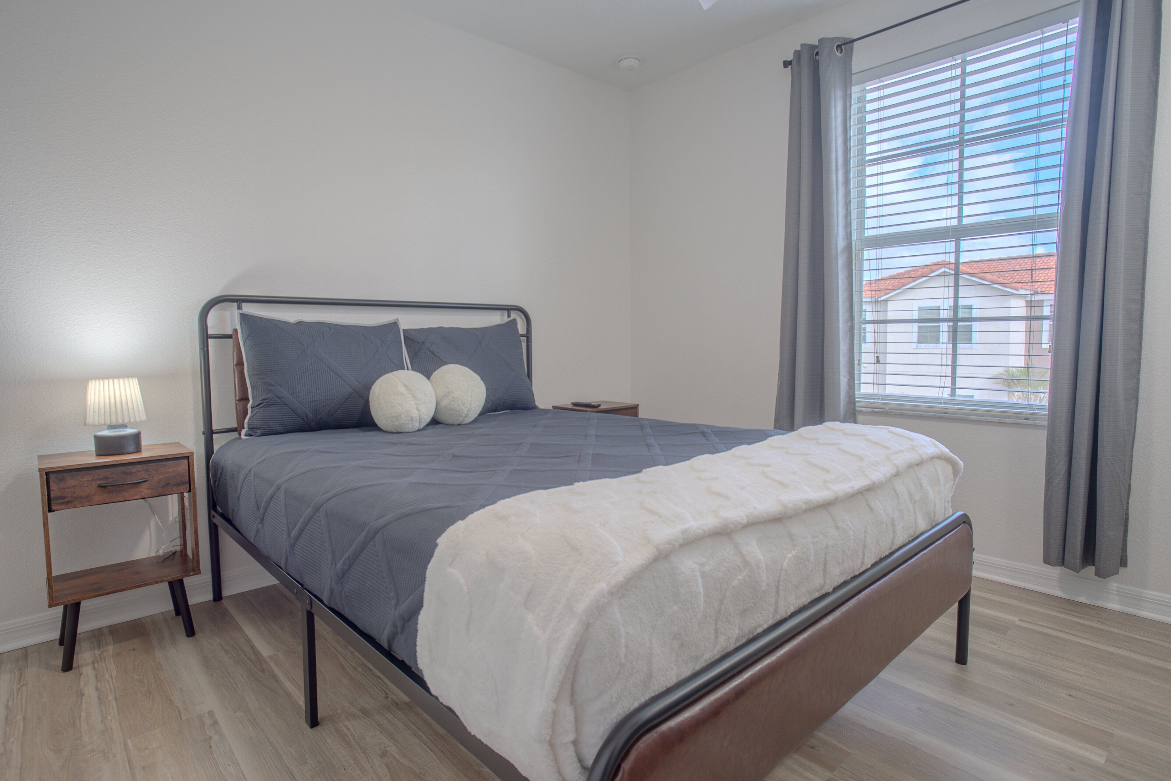A modern bedroom with a metal bed frame, a bedside table with a lamp, and a view of the outside through a window with blinds.