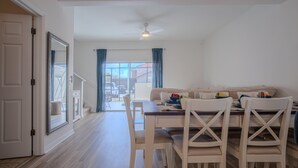 Un coin repas avec une table dressée pour quatre personnes, des chaises blanches, un ventilateur de plafond et une vue sur l’extérieur grâce à une grande fenêtre ornée de rideaux bleus.