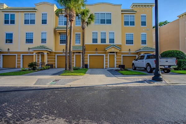 A multi-story residential building with a yellow facade, multiple garage doors, and a white pickup truck parked in front. - Spacious 3BR Townhouse in Vista Cay – Sleeps 8 Near Disney (Orlando)